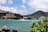 A sailboat is positioned on calm turquoise waters with a backdrop of green hills. In the background, there are buildings with red-tiled roofs and a larger cruise ship. The sky is partly cloudy, and nearby, green vegetation and rocky outcrops frame the scene on the edges.