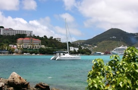 A sailboat is positioned on calm turquoise waters with a backdrop of green hills. In the background, there are buildings with red-tiled roofs and a larger cruise ship. The sky is partly cloudy, and nearby, green vegetation and rocky outcrops frame the scene on the edges.