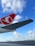 An airport scene featuring a large airplane wing with a red and white logo prominently displayed in the foreground. The sky is bright blue with scattered white clouds, creating a serene backdrop. In the distance, several other airplanes are positioned on the tarmac, along with a single safety cone visible on the ground.