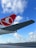 An airport scene featuring a large airplane wing with a red and white logo prominently displayed in the foreground. The sky is bright blue with scattered white clouds, creating a serene backdrop. In the distance, several other airplanes are positioned on the tarmac, along with a single safety cone visible on the ground.
