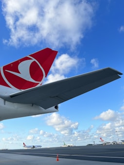 An airport scene featuring a large airplane wing with a red and white logo prominently displayed in the foreground. The sky is bright blue with scattered white clouds, creating a serene backdrop. In the distance, several other airplanes are positioned on the tarmac, along with a single safety cone visible on the ground.