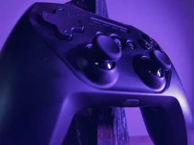A close-up of a black gaming controller illuminated by purple lighting. The controller features analog sticks, directional buttons, and various other buttons, highlighting its textured surface and design details.