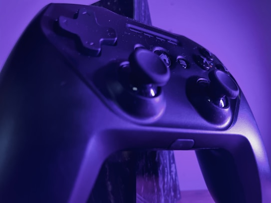 A close-up of a black gaming controller illuminated by purple lighting. The controller features analog sticks, directional buttons, and various other buttons, highlighting its textured surface and design details.