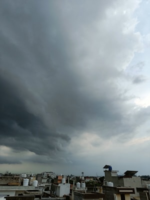 Dark, heavy clouds cover most of the sky, signaling an impending storm. Below, an urban landscape is visible with rooftops, water tanks, and buildings stretching into the distance.