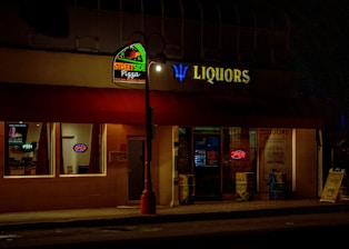 A dimly lit street scene featuring two adjacent storefronts, a pizza shop and a liquor store. The pizza shop has a brightly illuminated sign with 'StreetSide Pizza' and a neon 'OPEN' sign in the window. The liquor store displays a large 'LIQUORS' sign with an open illuminated door showcasing some bottles and products inside.