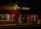 A dimly lit street scene featuring two adjacent storefronts, a pizza shop and a liquor store. The pizza shop has a brightly illuminated sign with 'StreetSide Pizza' and a neon 'OPEN' sign in the window. The liquor store displays a large 'LIQUORS' sign with an open illuminated door showcasing some bottles and products inside.