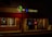 A dimly lit street scene featuring two adjacent storefronts, a pizza shop and a liquor store. The pizza shop has a brightly illuminated sign with 'StreetSide Pizza' and a neon 'OPEN' sign in the window. The liquor store displays a large 'LIQUORS' sign with an open illuminated door showcasing some bottles and products inside.