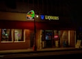 A dimly lit street scene featuring two adjacent storefronts, a pizza shop and a liquor store. The pizza shop has a brightly illuminated sign with 'StreetSide Pizza' and a neon 'OPEN' sign in the window. The liquor store displays a large 'LIQUORS' sign with an open illuminated door showcasing some bottles and products inside.