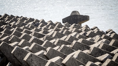 Geometric concrete structures are arranged in a repetitive pattern near a waterfront. The structures consist of interlocking blocks, forming a solid and uniform mass designed likely to serve as a breakwater or coastal defense. The background contains a body of water with a shimmering effect from the sunlight.