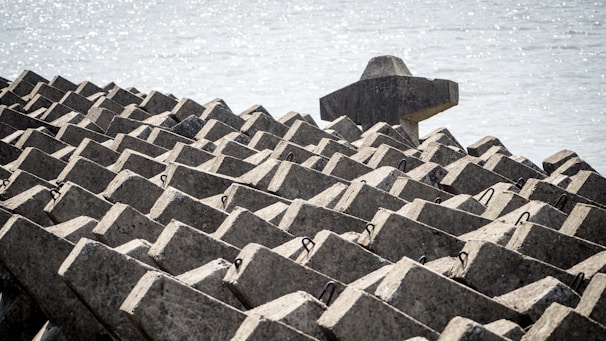Geometric concrete structures are arranged in a repetitive pattern near a waterfront. The structures consist of interlocking blocks, forming a solid and uniform mass designed likely to serve as a breakwater or coastal defense. The background contains a body of water with a shimmering effect from the sunlight.