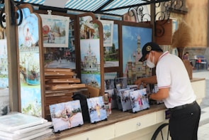 A person wearing a white shirt and cap is organizing a display of souvenir photos at an outdoor stand. The stand features large photos of attractions, possibly from an amusement park or similar venue. The photos are arranged on easels and there is a stack of cardboard boxes next to them. A striped canopy covers the stand, and there is a decorative plant on the counter.