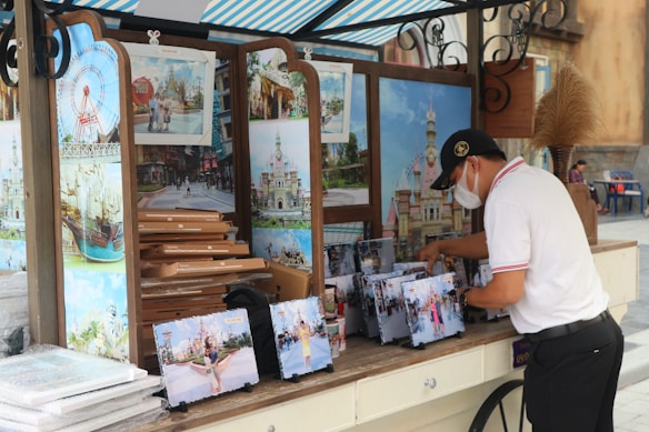A person wearing a white shirt and cap is organizing a display of souvenir photos at an outdoor stand. The stand features large photos of attractions, possibly from an amusement park or similar venue. The photos are arranged on easels and there is a stack of cardboard boxes next to them. A striped canopy covers the stand, and there is a decorative plant on the counter.