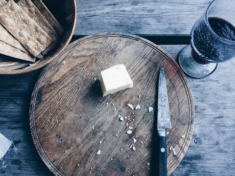 A rustic wooden table features a round wooden cutting board with a pat of butter. A Japanese knife with a black handle lays next to the butter on the board. Nearby, a bowl contains several rectangular sesame crackers. A glass of red wine sits off to the side.