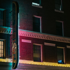 A hotel building exterior with a vintage neon sign at night, featuring glowing lights that cast a colorful glow onto the brick facade. The atmosphere is moody and nostalgic, with the architecture highlighting classic brickwork and framed windows.