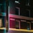 A hotel building exterior with a vintage neon sign at night, featuring glowing lights that cast a colorful glow onto the brick facade. The atmosphere is moody and nostalgic, with the architecture highlighting classic brickwork and framed windows.