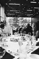 A formal event with people seated at elegantly set tables. Glasses, plates, and cutlery are neatly arranged. The setting appears to be indoors, with a large window in the background revealing trees outside. The ceiling is decorated with string lights, adding a warm ambiance.