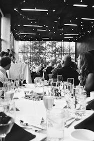 A formal event with people seated at elegantly set tables. Glasses, plates, and cutlery are neatly arranged. The setting appears to be indoors, with a large window in the background revealing trees outside. The ceiling is decorated with string lights, adding a warm ambiance.