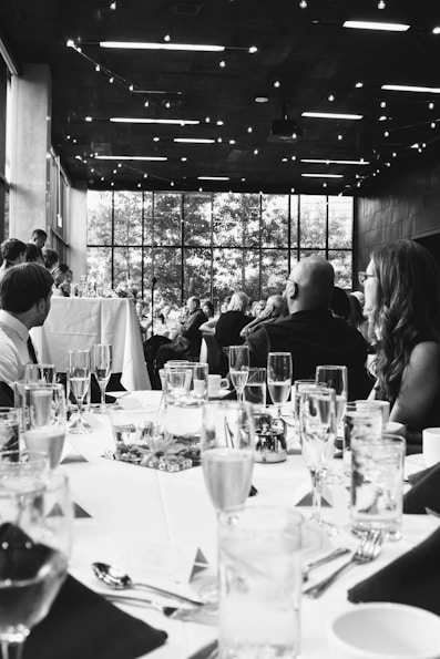 A formal event with people seated at elegantly set tables. Glasses, plates, and cutlery are neatly arranged. The setting appears to be indoors, with a large window in the background revealing trees outside. The ceiling is decorated with string lights, adding a warm ambiance.