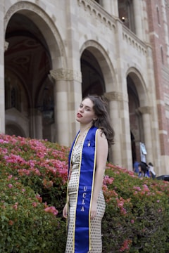 A woman in a patterned dress stands in front of an ornate building, wearing a blue graduation stole with 'UCLA' written on it. She is surrounded by vibrant pink flowers and lush greenery, with the architectural arches of the building visible behind her.