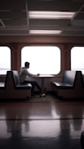 A person sits alone at a table in an empty ferry or train cabin, gazing out the large windows. The setting is serene with the interior featuring symmetrical seating arrangements and fluorescent ceiling lights.