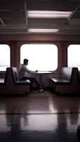 A person sits alone at a table in an empty ferry or train cabin, gazing out the large windows. The setting is serene with the interior featuring symmetrical seating arrangements and fluorescent ceiling lights.