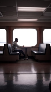 A person sits alone at a table in an empty ferry or train cabin, gazing out the large windows. The setting is serene with the interior featuring symmetrical seating arrangements and fluorescent ceiling lights.
