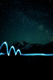 A night sky filled with numerous stars, featuring a backdrop of mountain silhouettes. A smooth, wavy line of blue light trails horizontally across the lower part of the image, suggesting motion or light painting.
