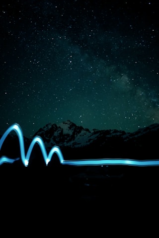 A night sky filled with numerous stars, featuring a backdrop of mountain silhouettes. A smooth, wavy line of blue light trails horizontally across the lower part of the image, suggesting motion or light painting.