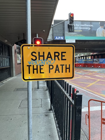 A yellow traffic sign with the words 'SHARE THE PATH' is mounted on a gray metal post. The sign has blue graffiti scribbled over it. Nearby, there is a black metal fence and a pedestrian sidewalk. Overhead, a traffic light shows a red light, and in the background, there’s a billboard that partially reads about recovery.
