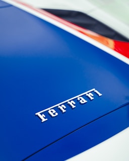 A close-up view of the Ferrari logo on the hood of a car, finished in a glossy blue color. The logo is centered and prominently displayed. The image captures a sleek and stylish design, with part of the car’s aerodynamic features visible in the background.