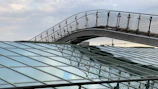 Metal and glass architectural structures with geometric patterns dominate the foreground. A pedestrian bridge with a grid-like railing arches over the scene. The background shows an industrial skyline, including cranes, under a cloudy sky.