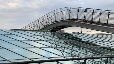 Metal and glass architectural structures with geometric patterns dominate the foreground. A pedestrian bridge with a grid-like railing arches over the scene. The background shows an industrial skyline, including cranes, under a cloudy sky.