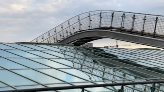 Metal and glass architectural structures with geometric patterns dominate the foreground. A pedestrian bridge with a grid-like railing arches over the scene. The background shows an industrial skyline, including cranes, under a cloudy sky.