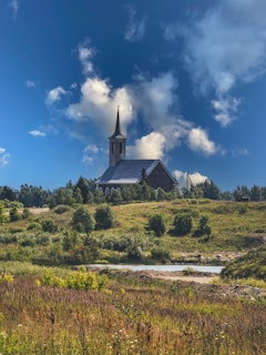 A serene countryside scene featuring a stone church with a tall steeple situated on a grassy hill. The sky is clear with scattered fluffy clouds, creating a peaceful backdrop. Surrounding the church are lush green bushes and trees, with a variety of flowering plants in the foreground.