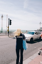A person with long blonde hair wearing a dark shirt with 'PHOTOGRAPHER' printed on the back is walking on a street near the seaside. They are holding a blue tote bag. A white car is driving on the road next to them. Lamp posts line the road, and the sea is visible in the background under a slightly overcast sky.