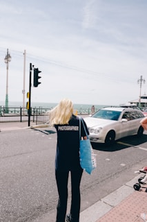 A person with long blonde hair wearing a dark shirt with 'PHOTOGRAPHER' printed on the back is walking on a street near the seaside. They are holding a blue tote bag. A white car is driving on the road next to them. Lamp posts line the road, and the sea is visible in the background under a slightly overcast sky.