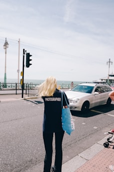 A person with long blonde hair wearing a dark shirt with 'PHOTOGRAPHER' printed on the back is walking on a street near the seaside. They are holding a blue tote bag. A white car is driving on the road next to them. Lamp posts line the road, and the sea is visible in the background under a slightly overcast sky.