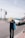A person with long blonde hair wearing a dark shirt with 'PHOTOGRAPHER' printed on the back is walking on a street near the seaside. They are holding a blue tote bag. A white car is driving on the road next to them. Lamp posts line the road, and the sea is visible in the background under a slightly overcast sky.