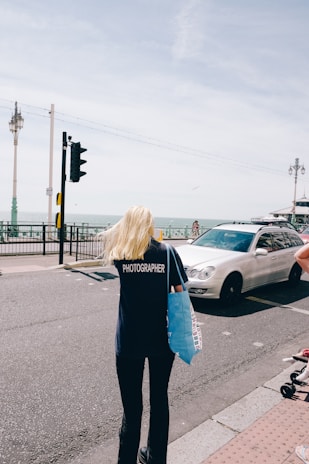 A person with long blonde hair wearing a dark shirt with 'PHOTOGRAPHER' printed on the back is walking on a street near the seaside. They are holding a blue tote bag. A white car is driving on the road next to them. Lamp posts line the road, and the sea is visible in the background under a slightly overcast sky.
