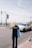 A person with long blonde hair wearing a dark shirt with 'PHOTOGRAPHER' printed on the back is walking on a street near the seaside. They are holding a blue tote bag. A white car is driving on the road next to them. Lamp posts line the road, and the sea is visible in the background under a slightly overcast sky.