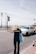 A person with long blonde hair wearing a dark shirt with 'PHOTOGRAPHER' printed on the back is walking on a street near the seaside. They are holding a blue tote bag. A white car is driving on the road next to them. Lamp posts line the road, and the sea is visible in the background under a slightly overcast sky.