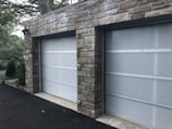A double garage with closed doors made of translucent panels framed by a stone structure. The garage is surrounded by a black asphalt driveway and some greenery is visible on the side.