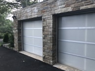 A double garage with closed doors made of translucent panels framed by a stone structure. The garage is surrounded by a black asphalt driveway and some greenery is visible on the side.