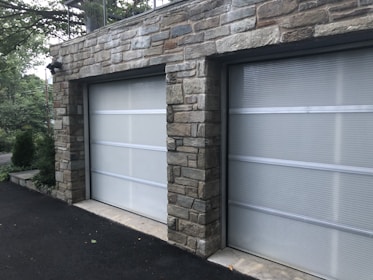 A double garage with closed doors made of translucent panels framed by a stone structure. The garage is surrounded by a black asphalt driveway and some greenery is visible on the side.