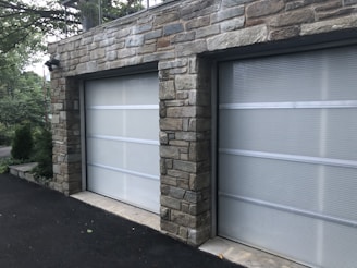 A double garage with closed doors made of translucent panels framed by a stone structure. The garage is surrounded by a black asphalt driveway and some greenery is visible on the side.