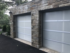 A double garage with closed doors made of translucent panels framed by a stone structure. The garage is surrounded by a black asphalt driveway and some greenery is visible on the side.