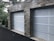 A double garage with closed doors made of translucent panels framed by a stone structure. The garage is surrounded by a black asphalt driveway and some greenery is visible on the side.