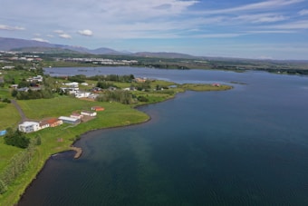 A picturesque lakeside scene featuring lush green fields, scattered homes, and a calm, expansive body of water under a lightly clouded blue sky. The landscape stretches into the distance with small hills and distant town structures visible.