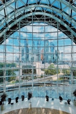 A large, curved glass wall dominates the foreground, offering a panoramic view of a modern city skyline in the background. The glass structure features an intricate metal framework. Inside the building, several chairs and potted plants are arranged along the curve, suggesting a spacious, indoor viewing area. The blue sky and reflection of skyscrapers create a sense of openness and connectivity with the urban landscape.