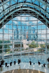 A large, curved glass wall dominates the foreground, offering a panoramic view of a modern city skyline in the background. The glass structure features an intricate metal framework. Inside the building, several chairs and potted plants are arranged along the curve, suggesting a spacious, indoor viewing area. The blue sky and reflection of skyscrapers create a sense of openness and connectivity with the urban landscape.